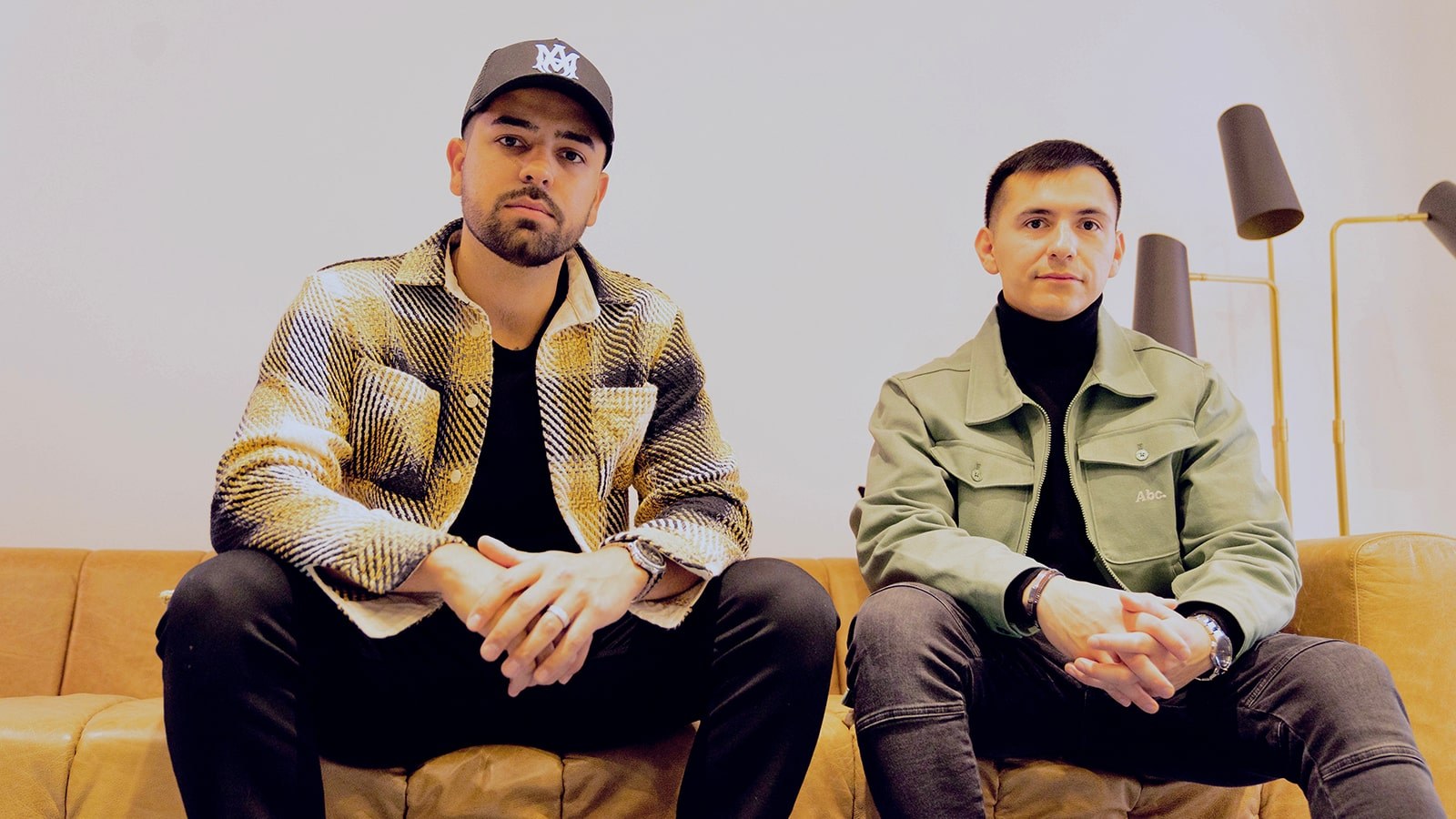 Two men seated side by side on a light brown leather couch against a white backdrop.