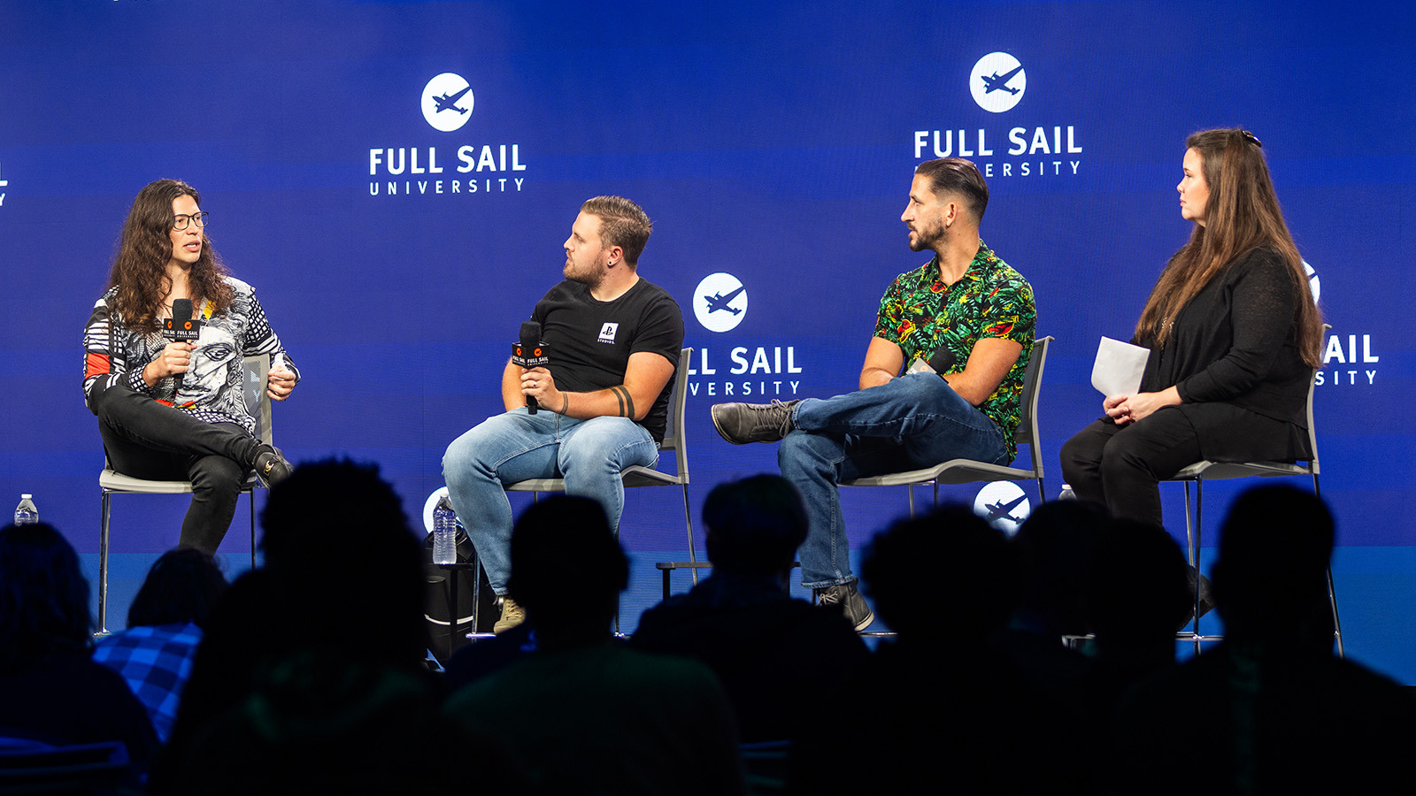 Four people seated on a stage, talking and holding microphones in front of a large crowd.