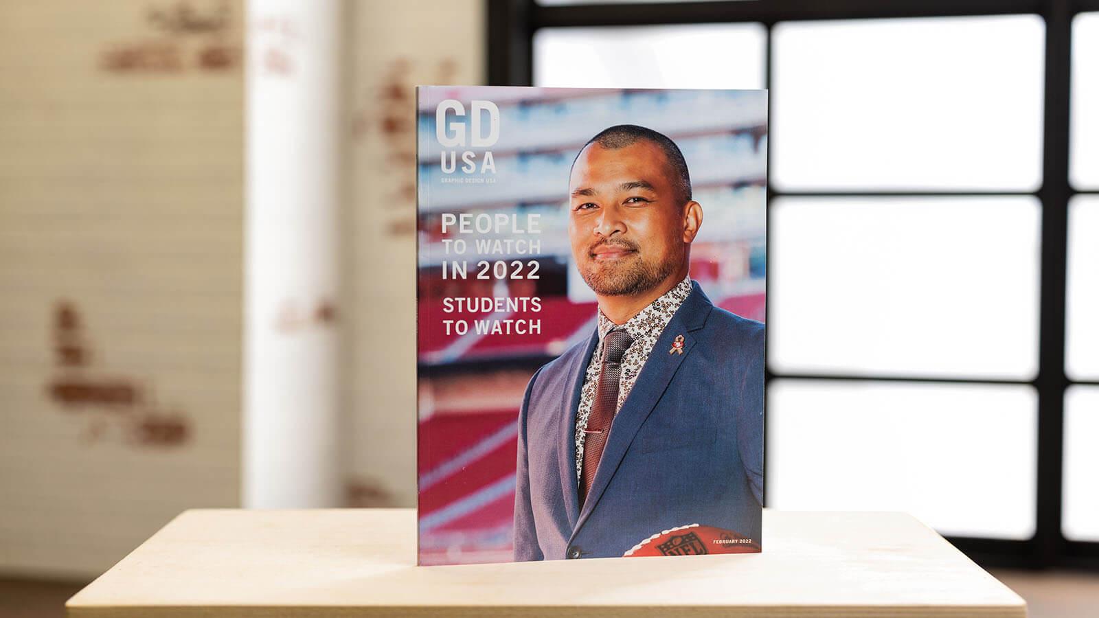 A Graphic Design USA magazine stands upright on a desk in a backlit room. On the cover, a man wearing a blue suit with a pink ribbon pin and holding an NFL football smiles for the camera. The cover reads, People to Watch in 2022, Students to Watch.