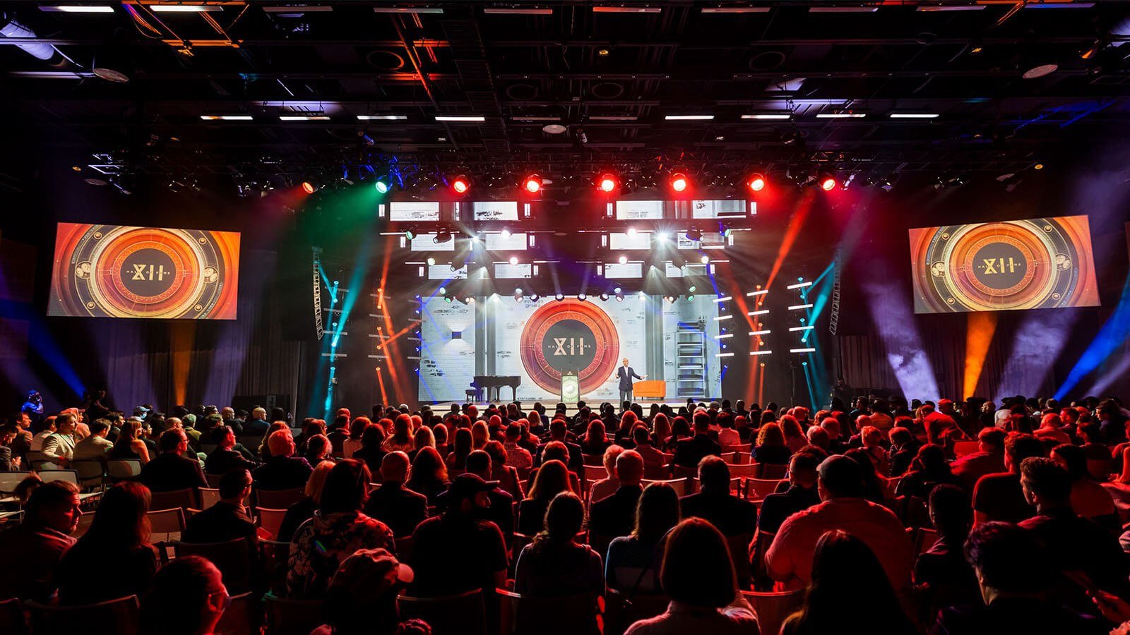 A person wearing a suit speaks on a stage in front of a large crowd. There is a large screen with a logo that says Hall of Fame XII behind them. Red, blue, and white lights illuminate the room.