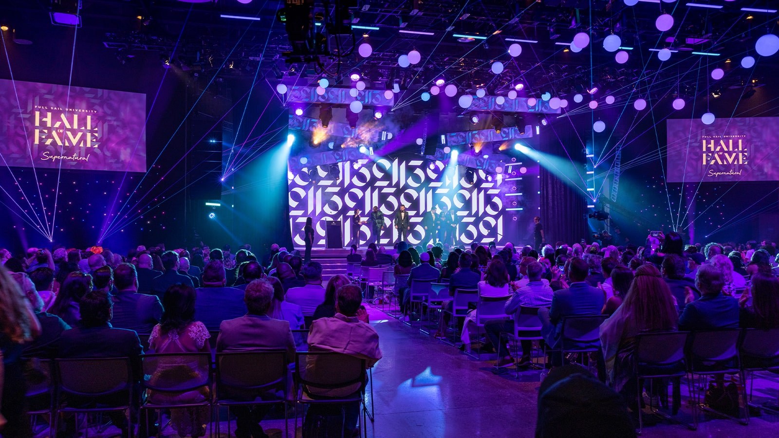 The Hall of Fame 2023 inductees stand on a stage, surrounded by beautiful purple lighting in a large auditorium. The audience is applauding.