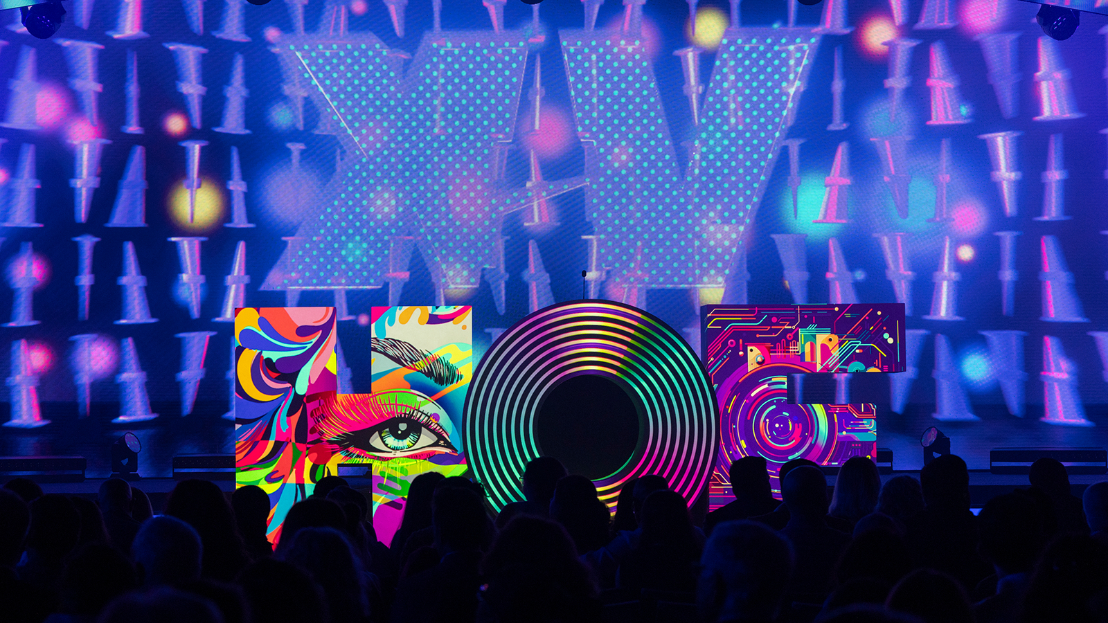 A large sculpture of the letters “HOF” sits atop the Full Sail Live Venue stage with different multicolored images projected onto each letter. The letters are in front of a large purple screen featuring the Roman numeral “XV” in large font. In front of the stage is a black silhouette of the crowd.