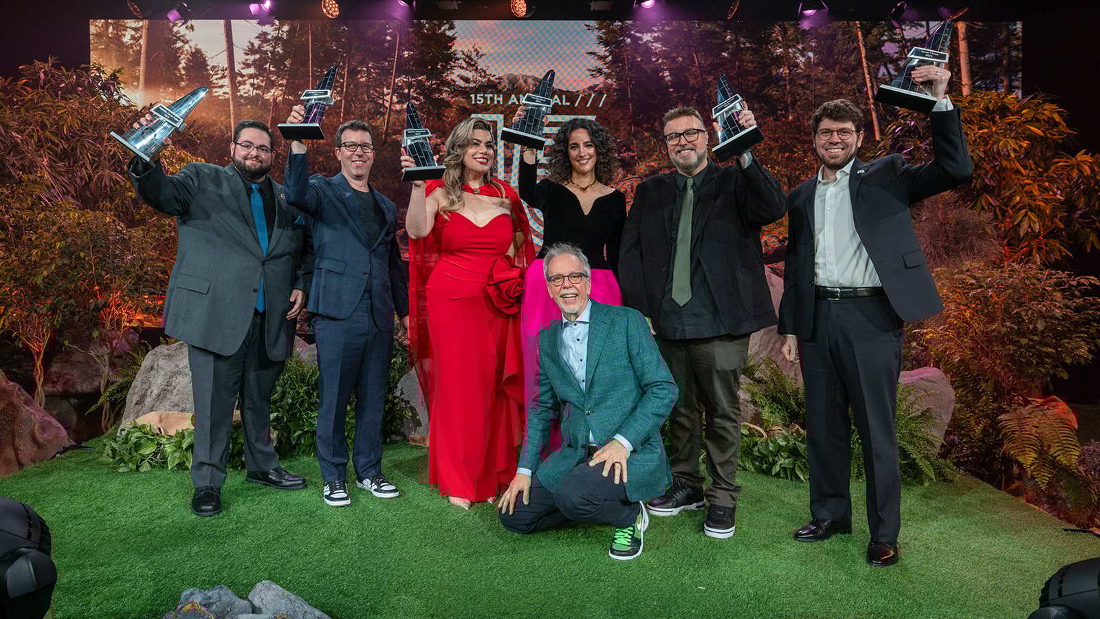 The Hall of Fame 15 inductees raise their awards in the air on a stage decorated with foliage. Garry Jones kneels in front.