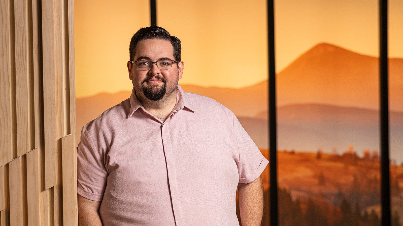 A man with glasses and a beard in a pink button-down stands next to a wooden panel, with a warm, mountainous sunset background behind him.