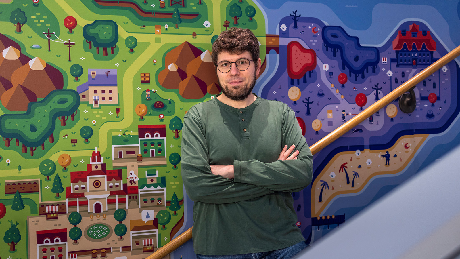 A bearded man in glasses and a green shirt stands with his arms crossed in front of a colorful, game-inspired illustrated mural.