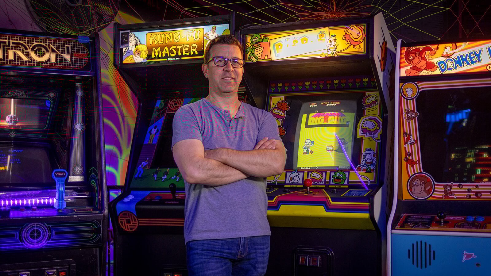 Jonathan Mayer smiles and stands in front of a row of arcade game machines for ‘Tron’, ‘Kung-Fu Master’, ‘Dig Dug’, and ‘Donkey Kong’.