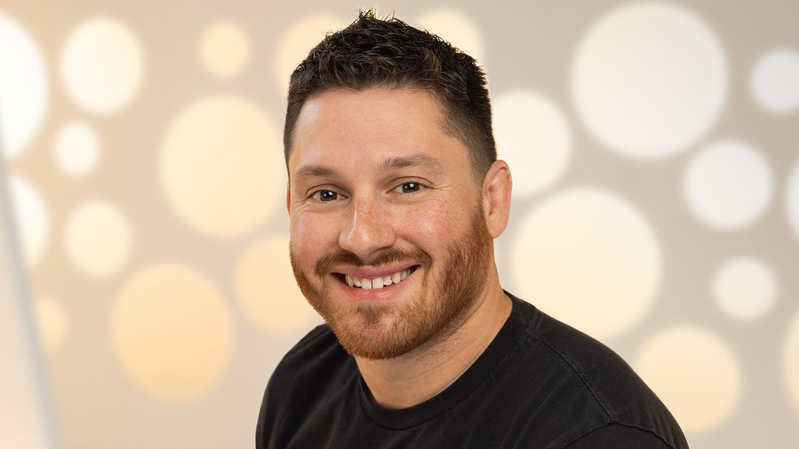 A headshot of a man with short brown hair and a black t-shirt smiling against a white background.