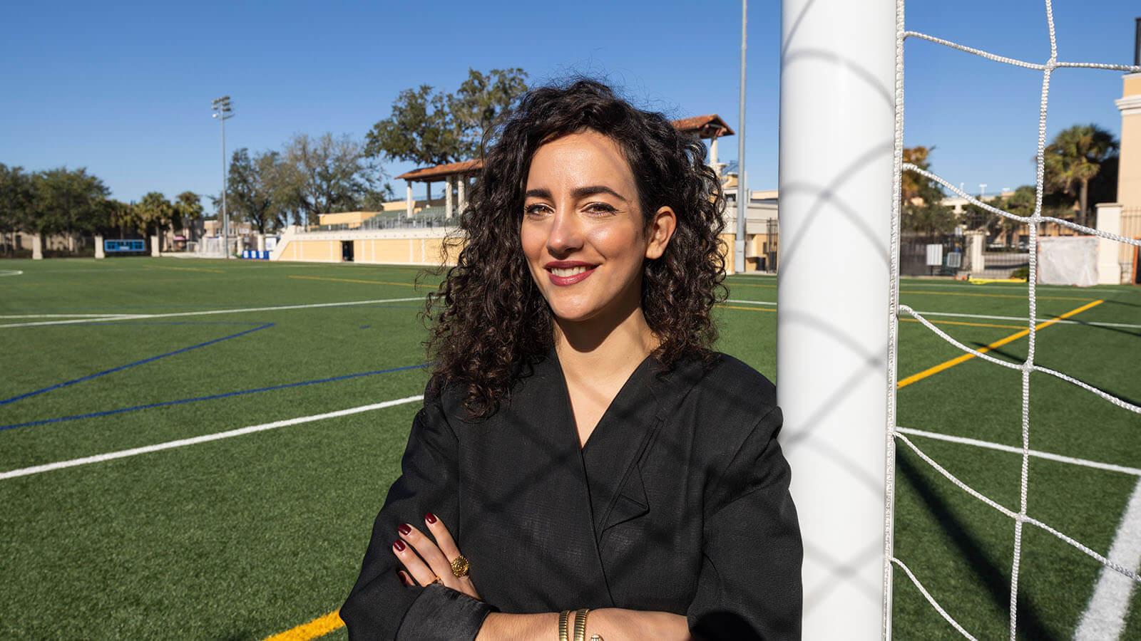 A woman with curly hair and a black jacket stands confidently by a soccer goalpost on a bright, sunny day at an outdoor field.