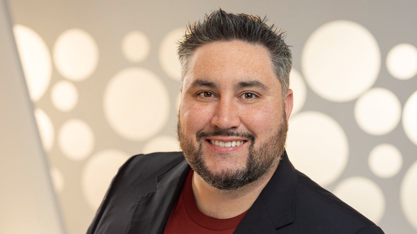 Michael La Plante smiles at the camera. He is wearing a red t-shirt with a black blazer and standing in front of a white backdrop.