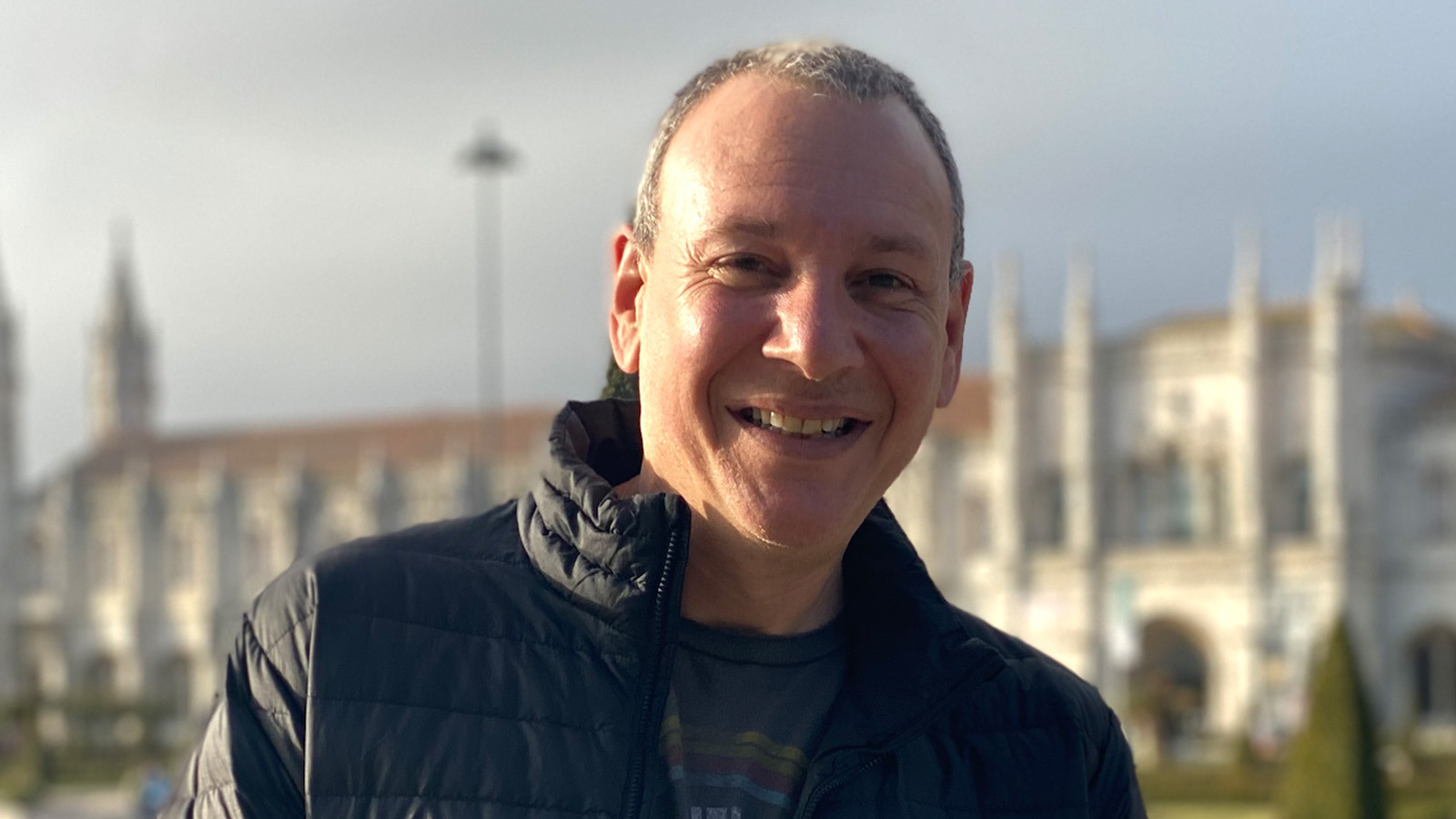 Sean Spuehler stands outdoors in front of a large building. He is smiling and wearing a black jacket and a blue t-shirt.