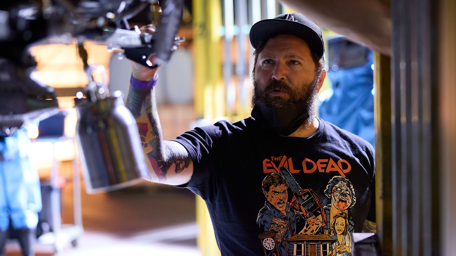 Steven C. Miller adjusts a piece of machinery on a science lab set. He is wearing an Evil Dead t-shirt.