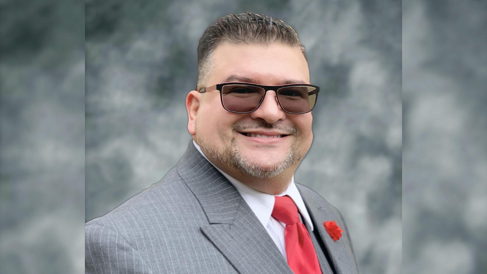 Will Torres wears a gray pinstriped suit with a red tie, a red carnation on the lapel, and sunglasses.