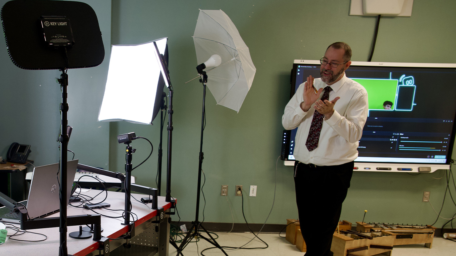 An instructor gestures while teaching in a studio setup with lights, cameras, and a green screen projection in the background.