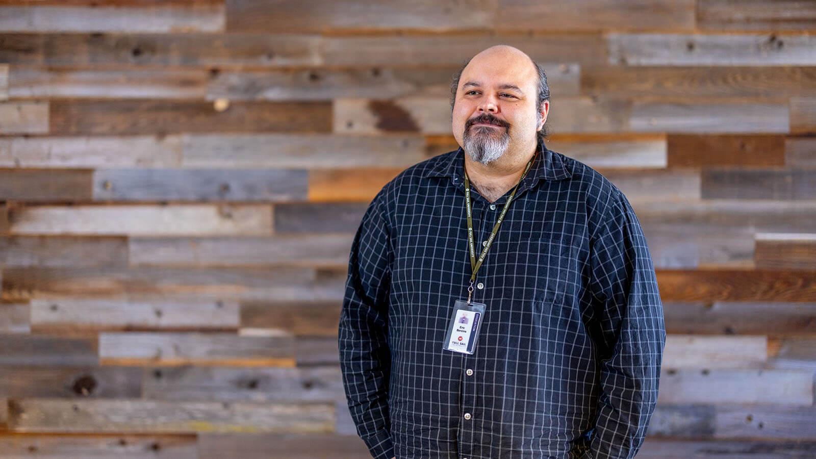 Instructor Eric Berzins, a man with a grey goatee, standing in front of a wood paneled wall wearing a blue button down dress shirt and green Full Sail lanyard with name badge.
