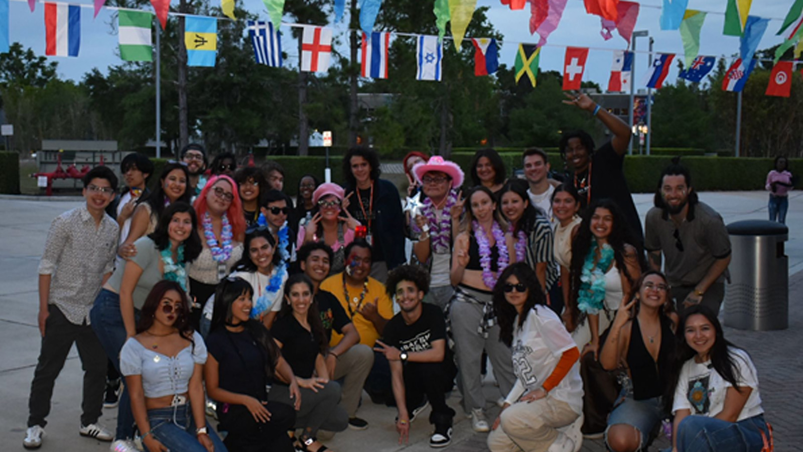 A diverse group of young adults poses outdoors under international flags, dressed festively with leis and fun accessories.