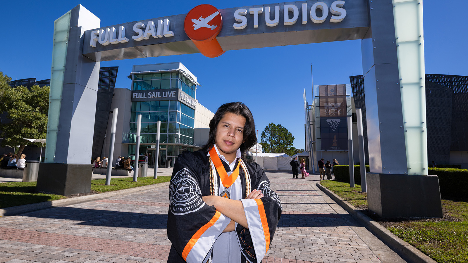 John Tobar stands with his arms crossed in front of the Full Sail Studios archway. He is wearing a graduation gown and an Armada medallion.