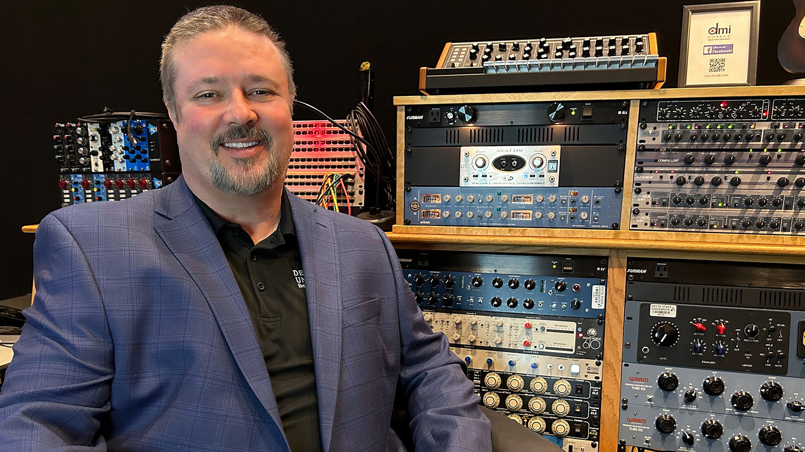 Richard Tremmel smiles at the camera with an array of music recording equipment behind him. He is wearing a black shirt and blue blazer.