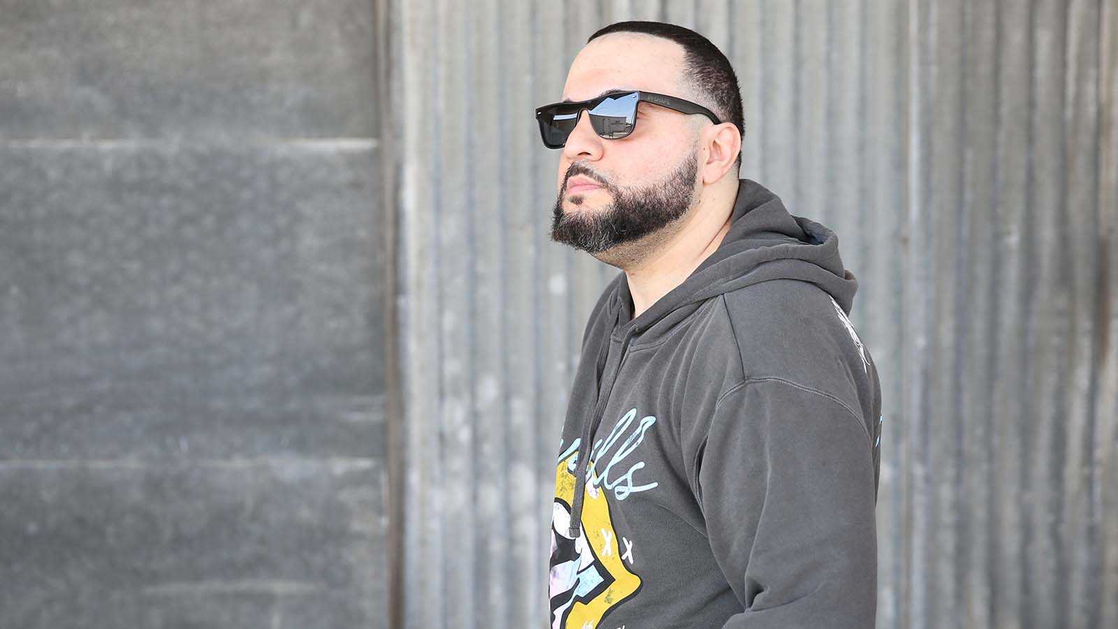 Full Sail grad George Doman wearing black sunglasses and a grey pullover hoodie with a yellow and light blue design, standing in front of a large stone wall while looking off into the distance.