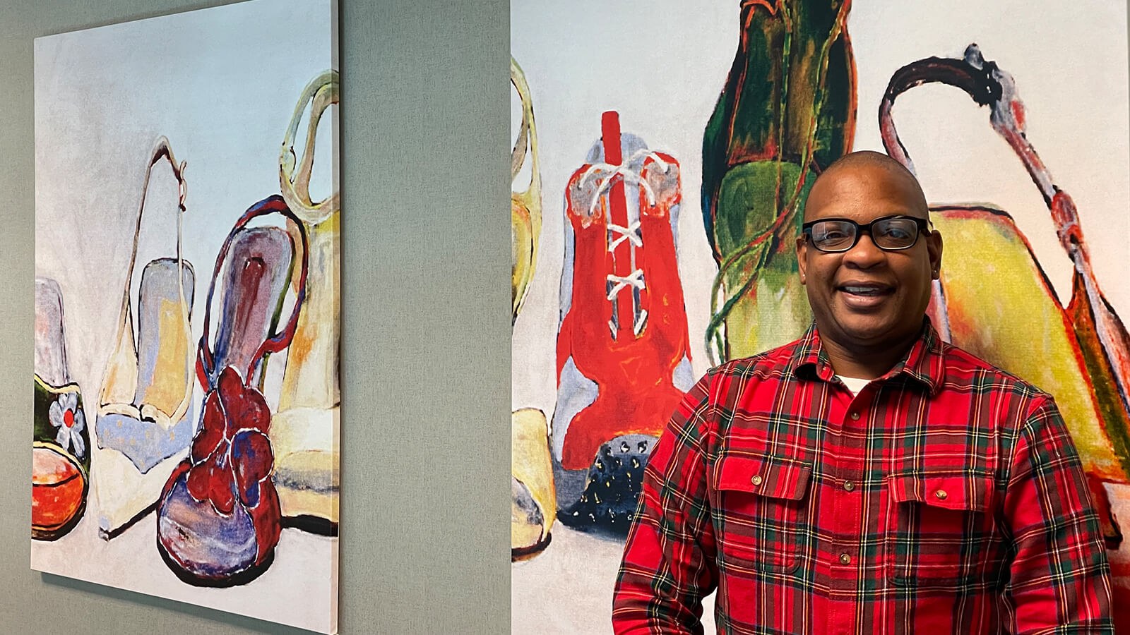 Anthony Roberts Jr. stands in front of two paintings of shoes. He is wearing black-rimmed glasses and a plaid button-down shirt.