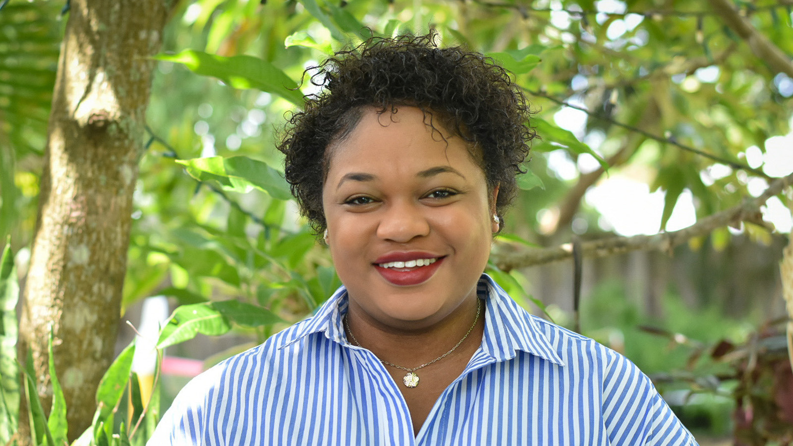 Jessica stands in front of a lush garden, surrounded by greenery. She is wearing a blue pinstripe shirt and is smiling.