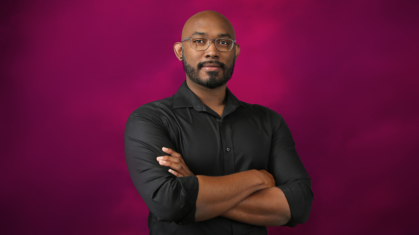 Gregg is wearing a black shirt and rectangular glasses. He is standing in front of a pink background with his arms crossed.