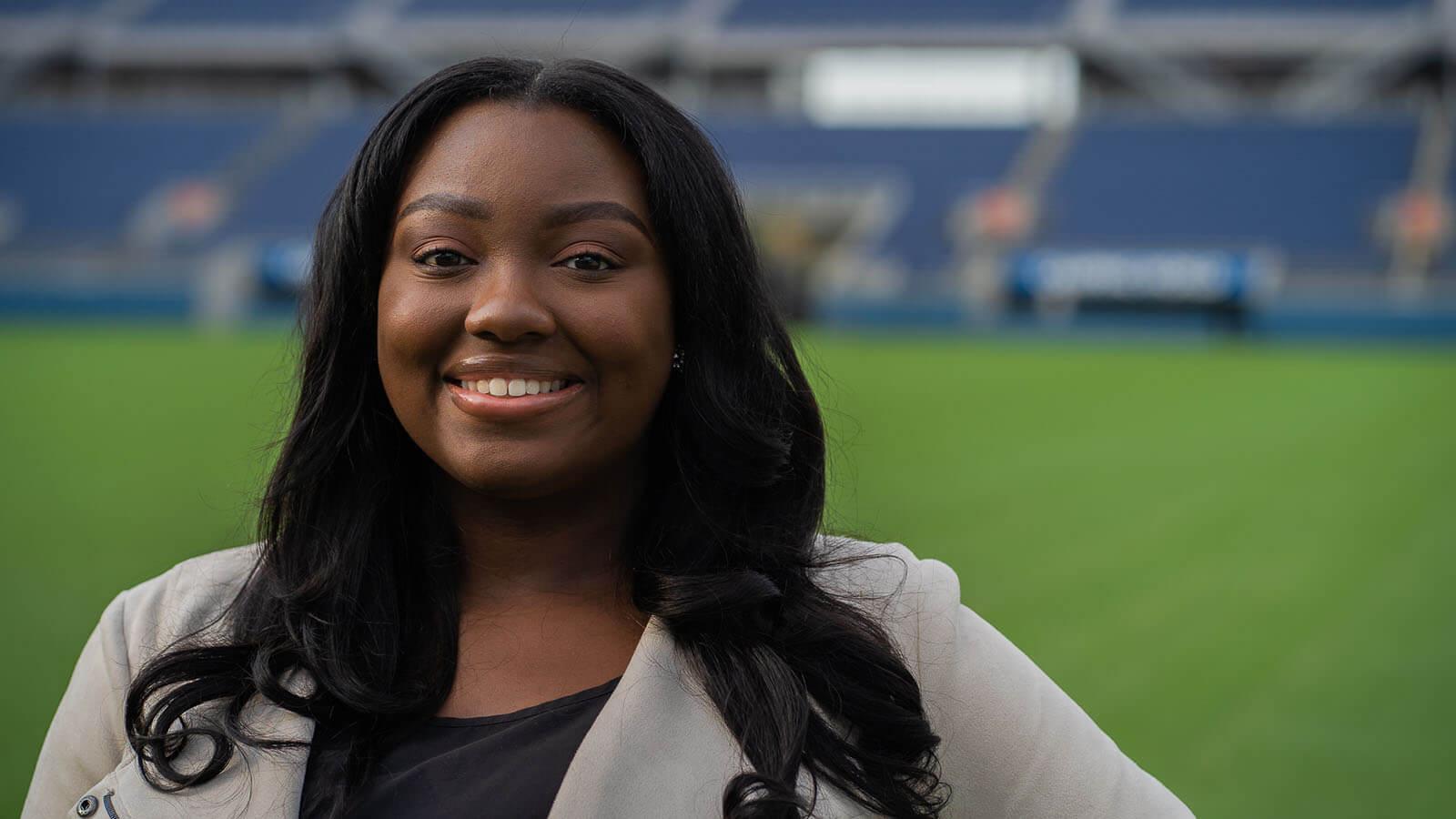 A woman with long brown hair smiling in a beige blazer while standing on a soccer pitch.