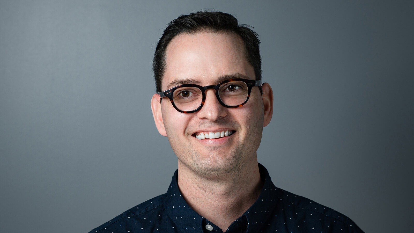 Digital Arts & Design grad Paul Shank wearing a blue, polka-dotted shirt in front of a gray background.