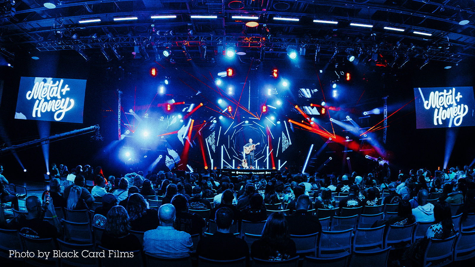 A vibrant charity concert featuring Matt Heafy of Trivium on stage with dynamic lights, a packed audience, and 'Metal + Honey' branding on screen.