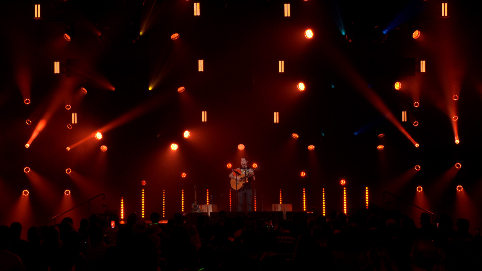 Trivium lead-singer Matt Heafy on a dimly-lit stage playing an acoustic guitar in front of a large crowd.