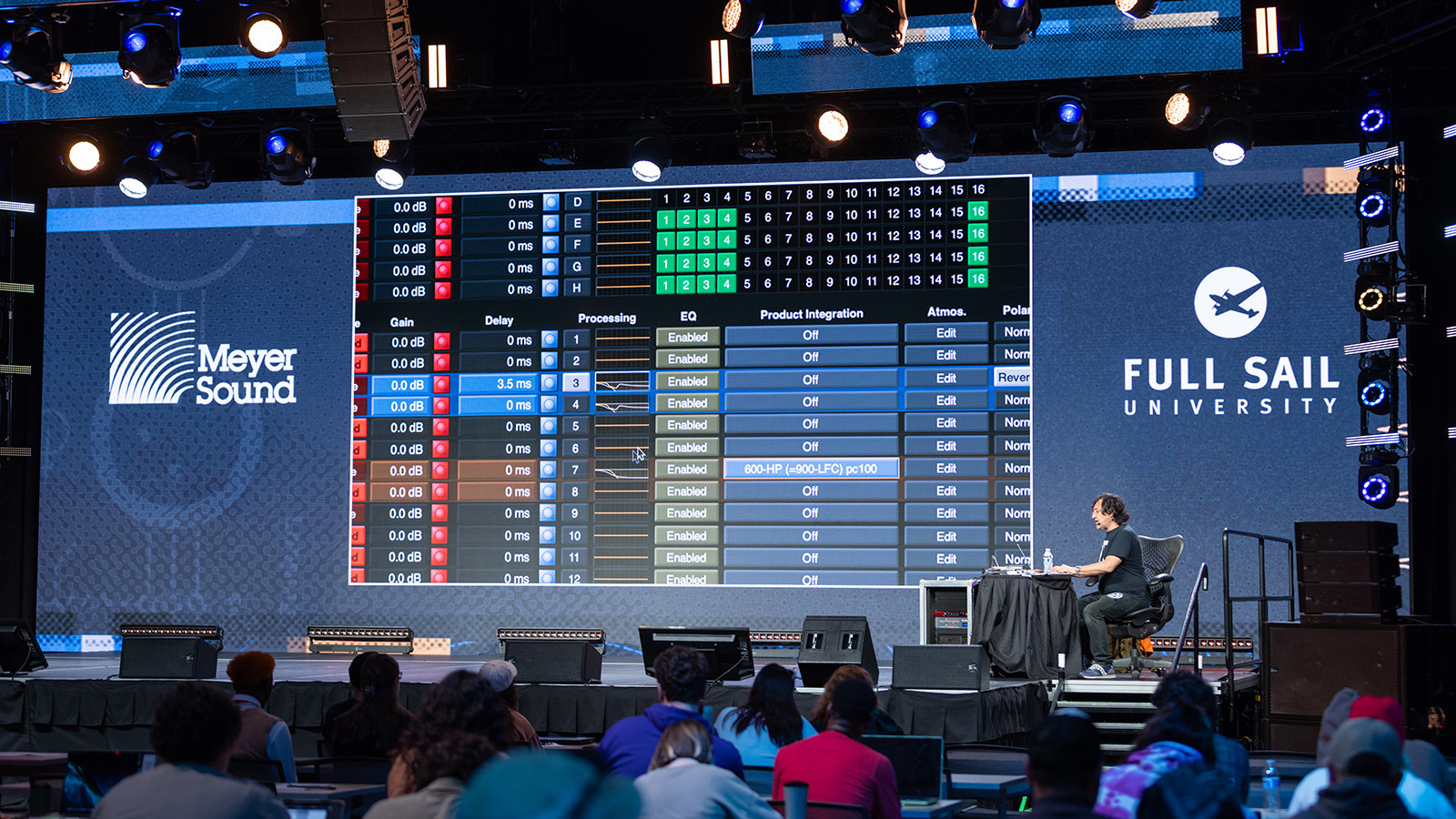 A person is seated at a desk on a stage in front of a crowd of students while a large LED screen behind displays a mirror of the audio program running on their computer.