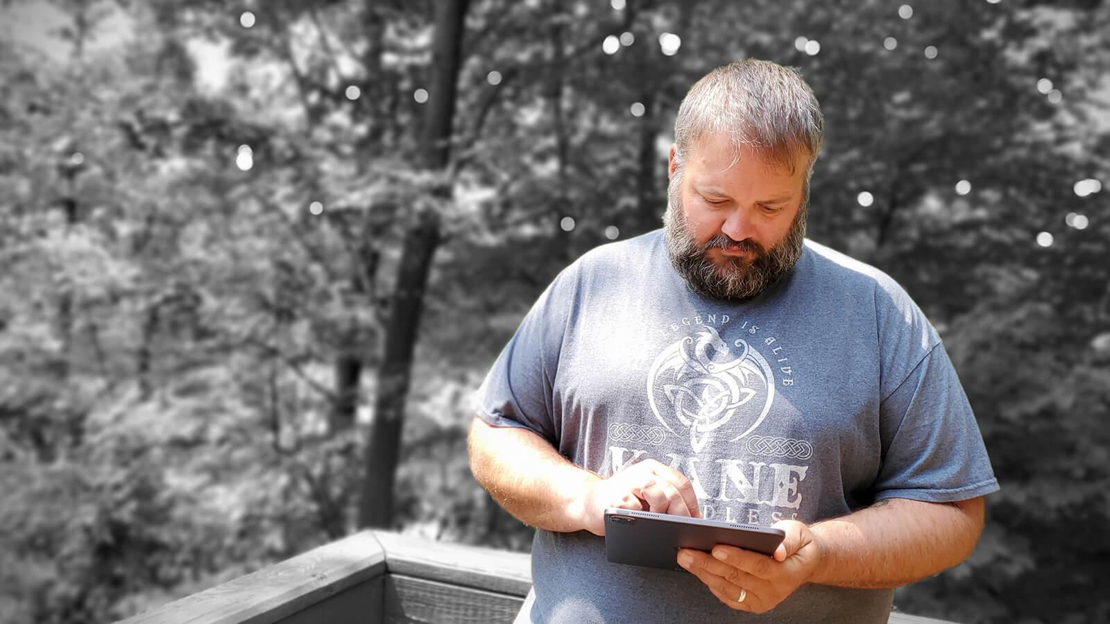 Grad Ryan Backa in a blue shirt with a white dragon design, holding an electronic tablet in front of a wooded area in greyscale.