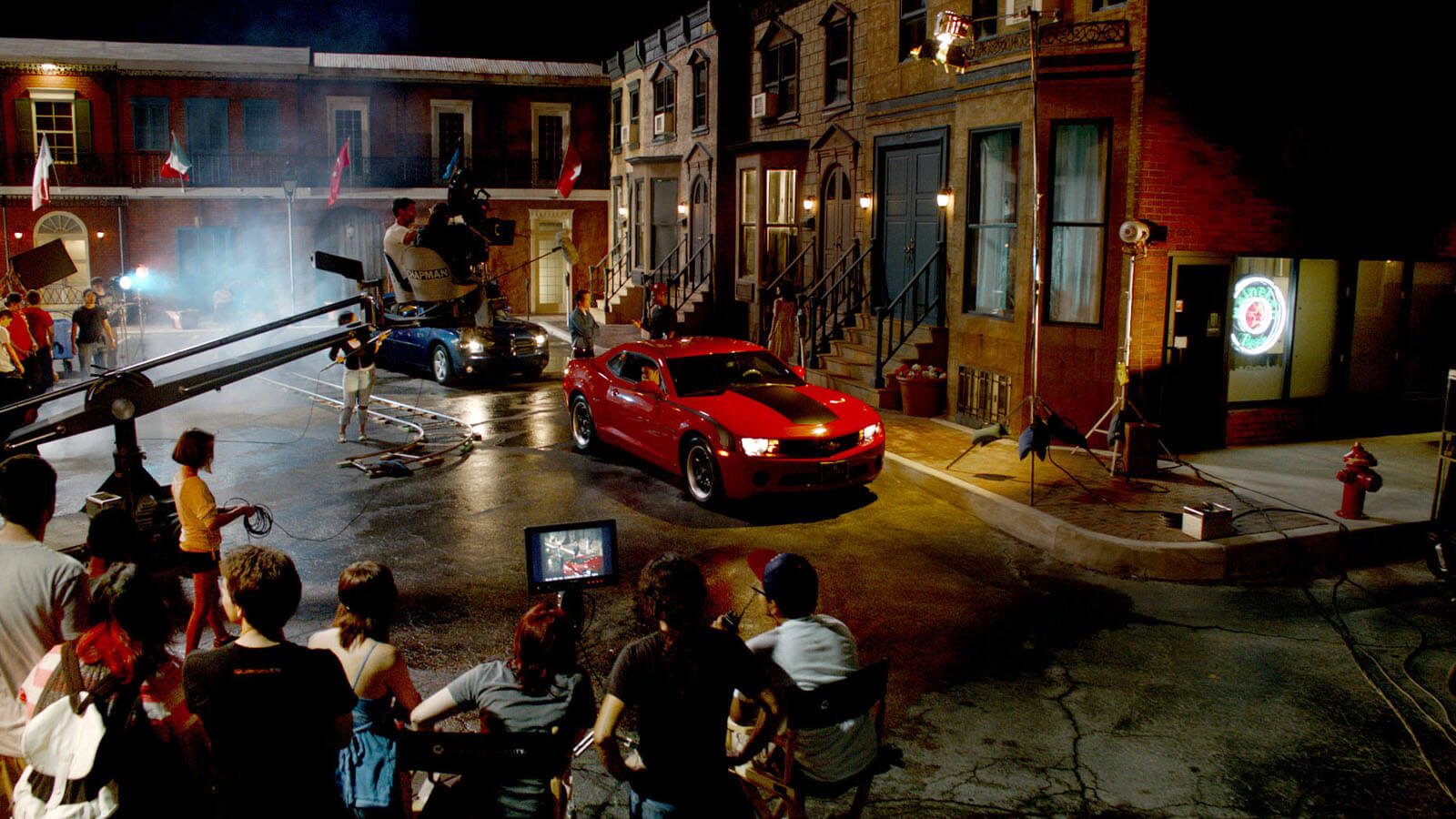 Full Sail University's Backlot in the evening filled with a full film production crew shooting a scene featuring two muscle cars, one red and one black.