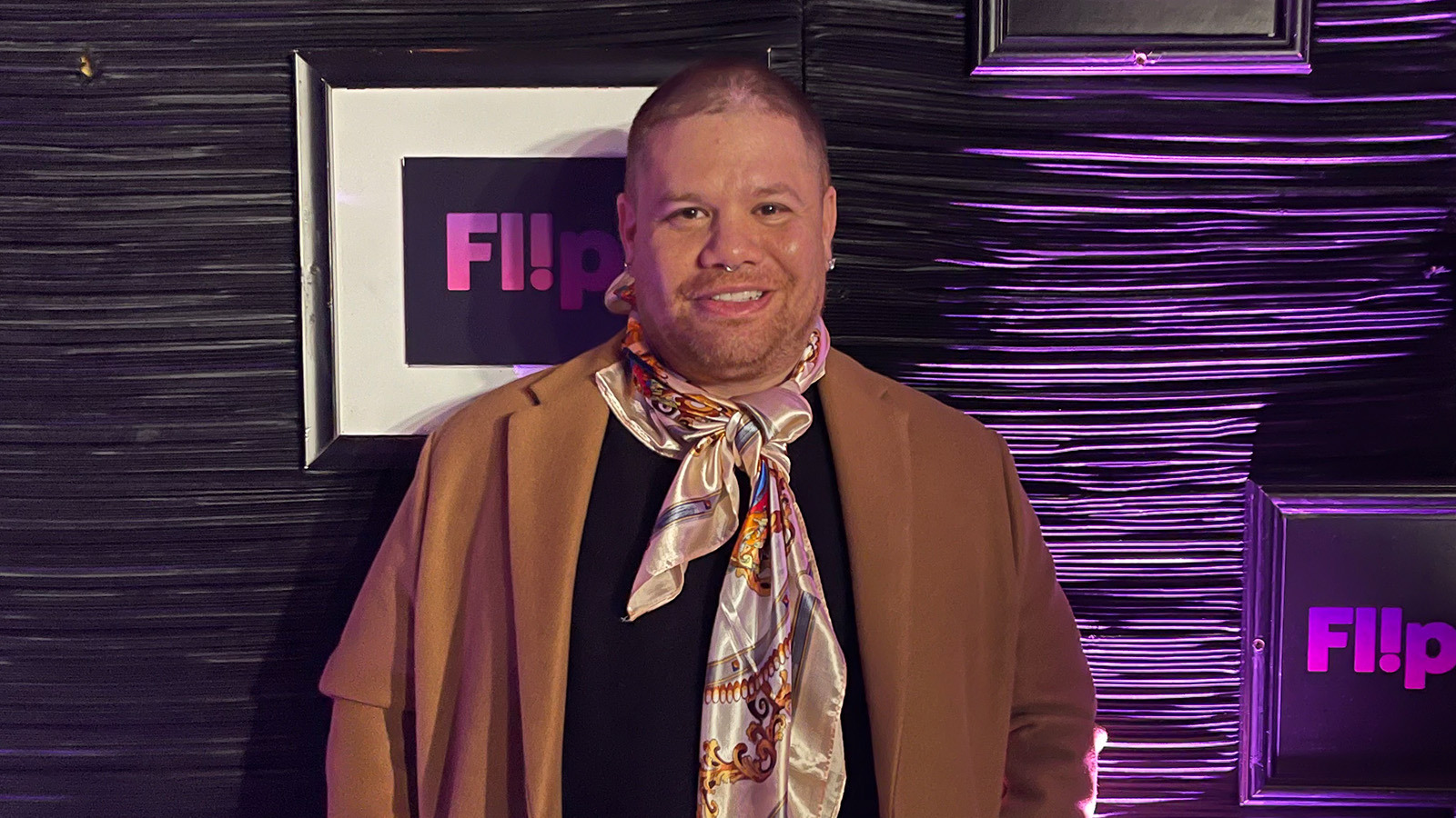 Javi Simons stands in front of a purple wall. He wears a black t-shirt, jeans, a brown overcoat, and a colorful scarf. He is smiling.