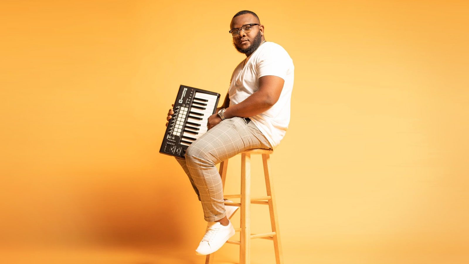 Chavez Parker sits on a stool and holds a keyboard. He wears glasses, a t-shirt, brown pants, and sneakers.