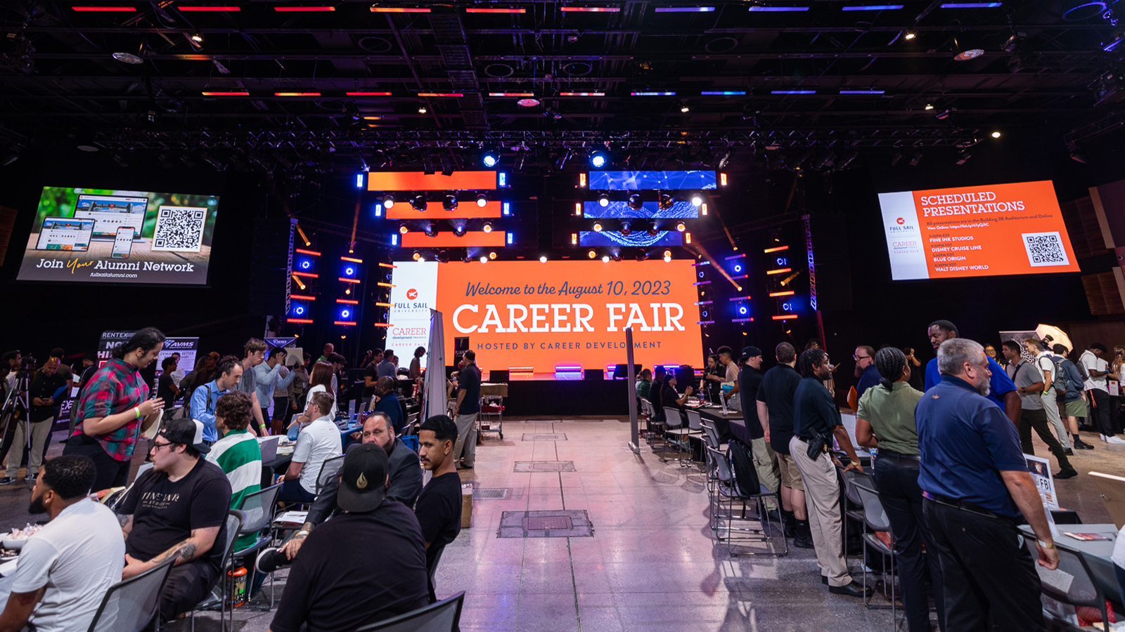 A bustling career fair with several booths set up. In the center, a giant LED screen welcomes guests to the Career Fair.