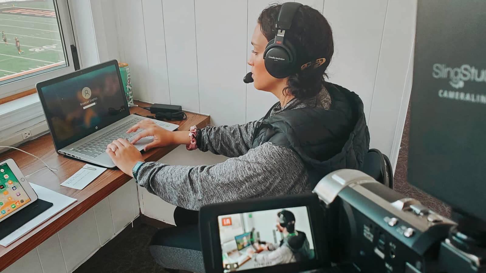 Grad Sydney Phillips, a woman with dark brown hair wearing a grey hooded sweatshirt and black over-ear headphones, is seated at a desk on her laptop in a press box overlooking athletes in uniform on a football field.