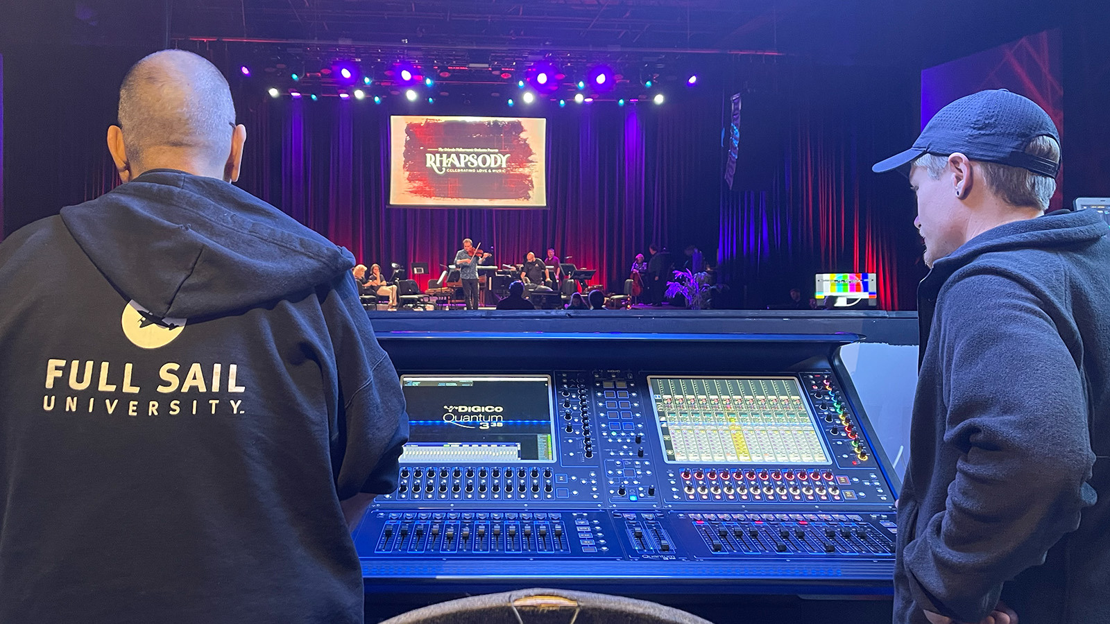Two people's backs are facing the camera as they overlook an audio console and stage at The Plaza Live.