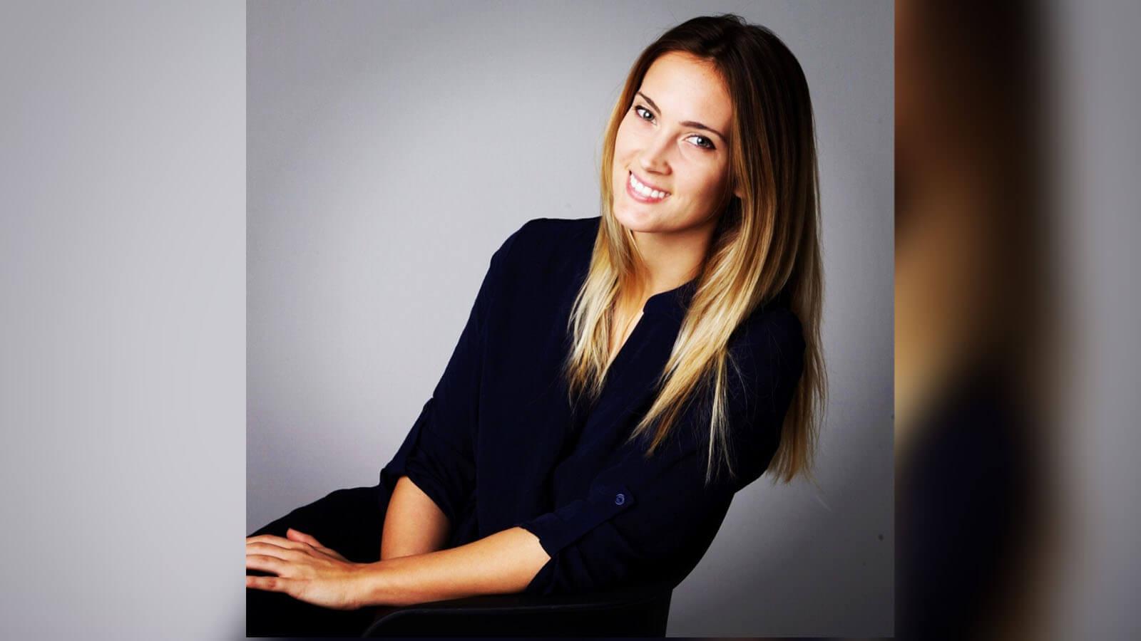 Grad Lisa Maskey, a smiling woman with straight blond hair wearing a black three-quarter sleeve top seated against a grey backdrop.