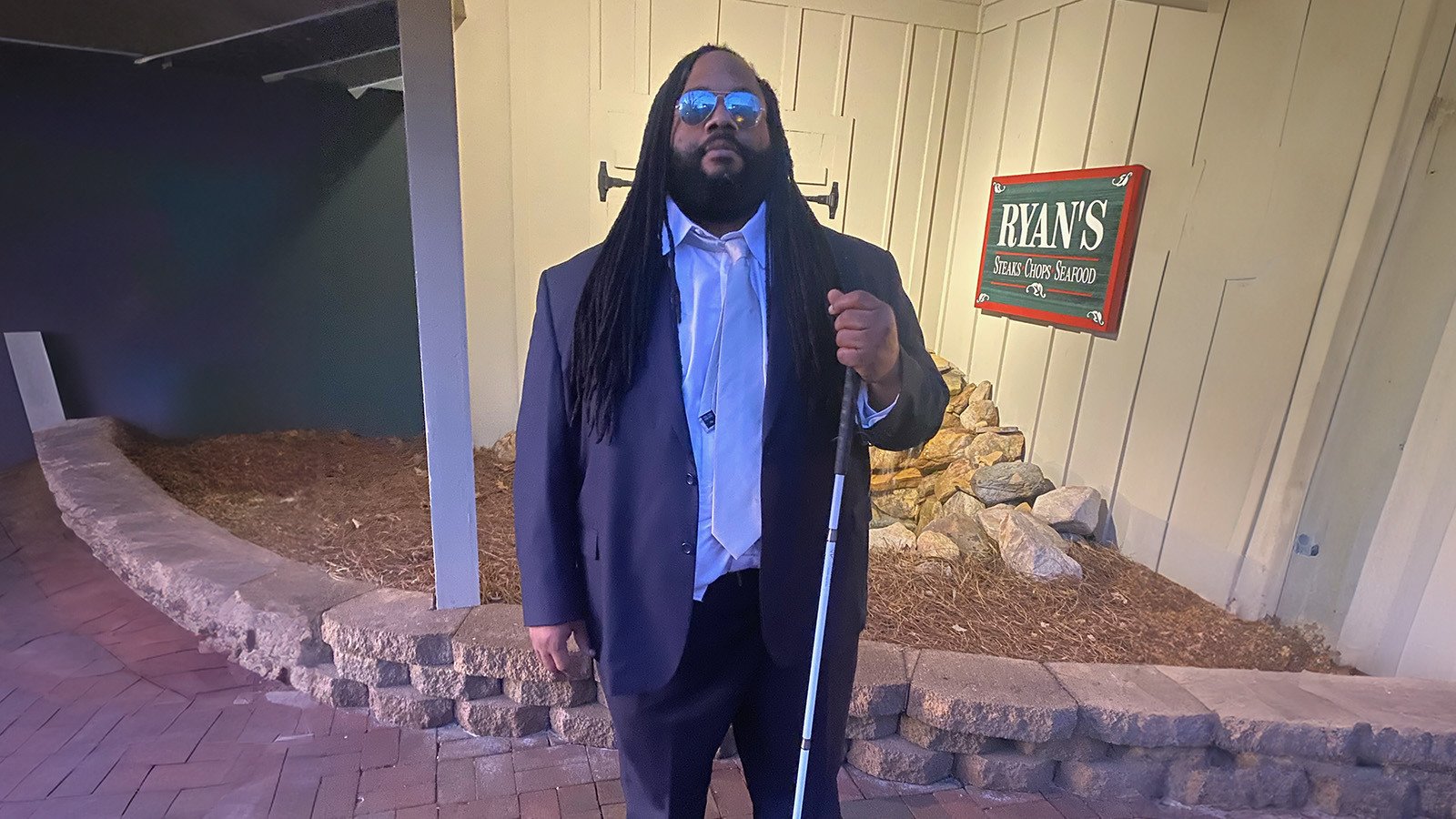 Andre Woods stands outdoors in front of a restaurant. He is wearing a blue suit and sunglasses. He is holding a mobility cane.