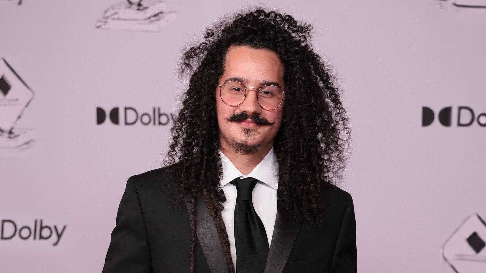 Guillermo Moya smiles at the camera in front of a white background. He is wearing a black suit, black tie, and glasses.