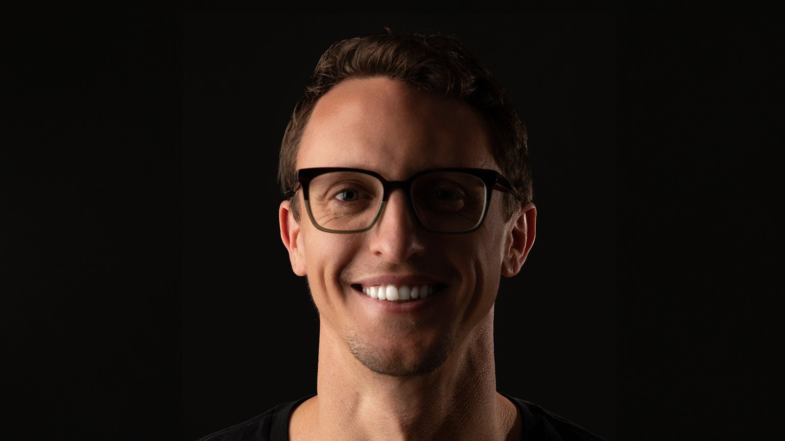 Drew Becker smiles at the camera in front of a dark background. He is wearing black glasses and a black shirt.