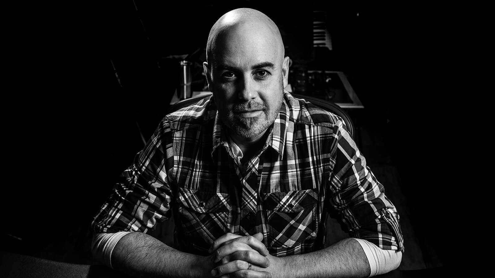 Grad Terry Watson sits with his hands clasped at a desk. He is wearing a plaid button-down shirt and is in a music studio.