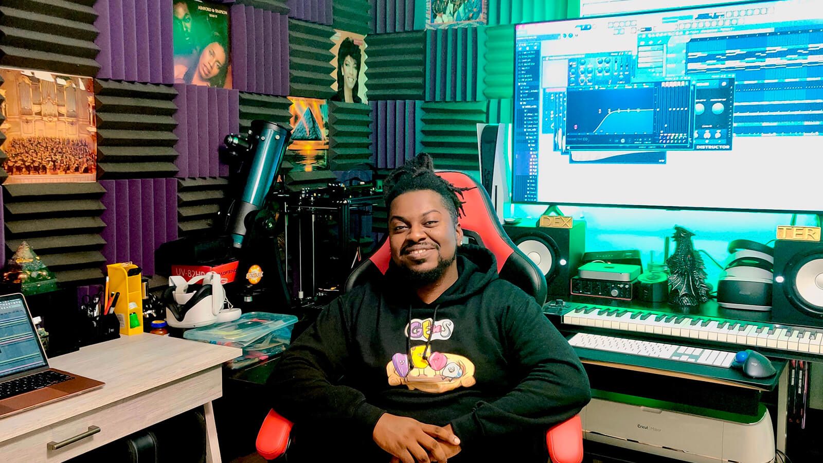 Grad Shawn Deshield smiling while seated at a desk in a home studio featuring a keyboard, audio equipment, and a large screen displaying audio software while wearing a black hooded sweatshirt with a colorful design.