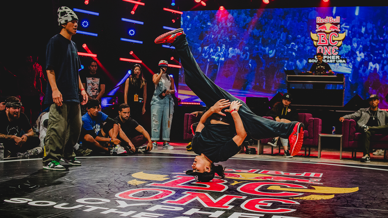 A dancer is doing a hands-free headstand with their legs spread while their competitor, the audience, and the judges watch.