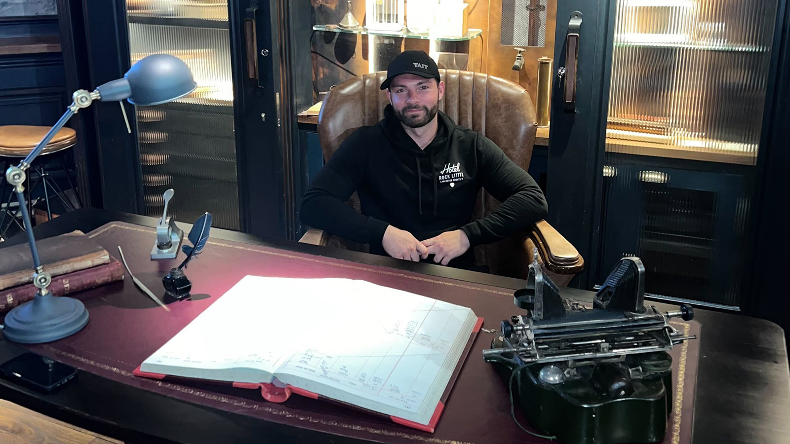 Rob Prager sits in a leather chair behind a large desk. He is smiling and wearing a black baseball cap and a black hoodie.