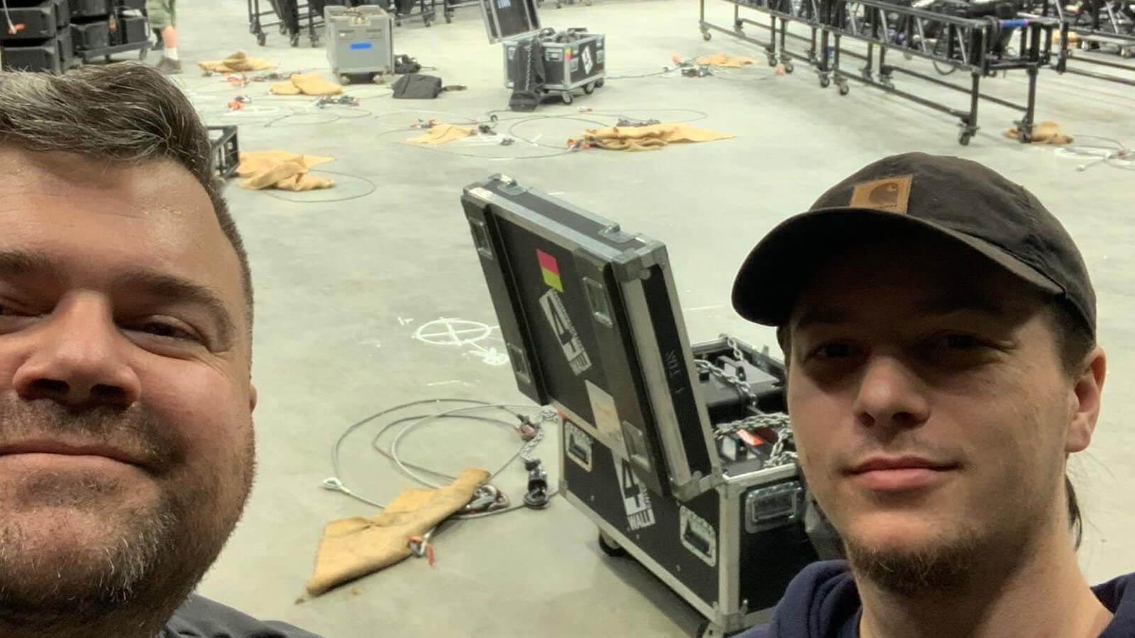 Two smiling men with beards stand in a backstage area. There are wires on the floor behind them, as well as an open trunk filled with production equipment.