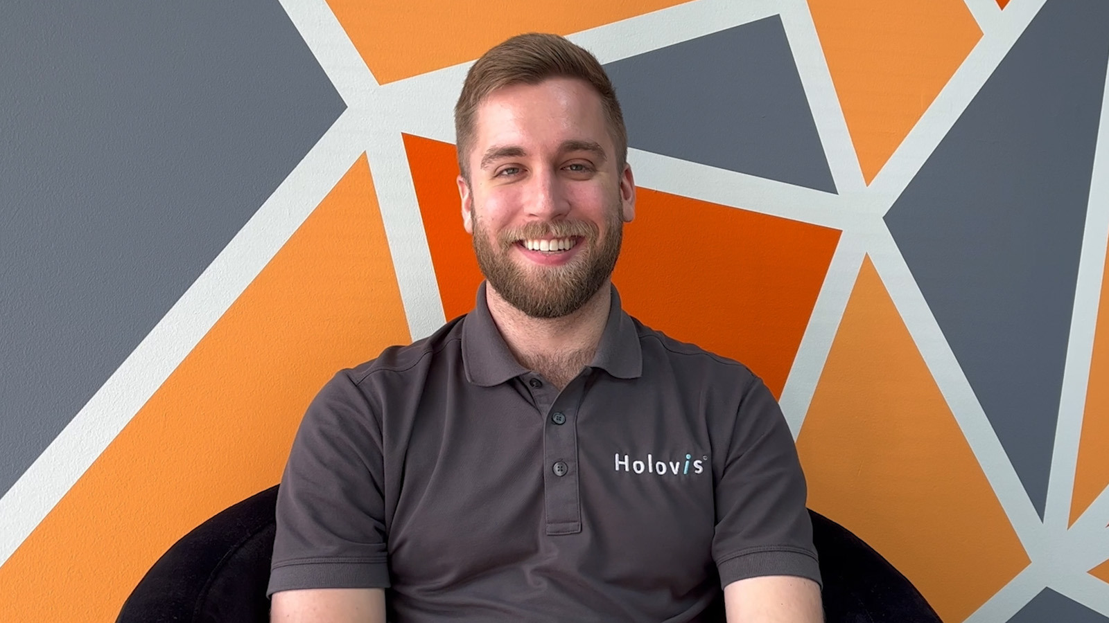 A man with light hair and a beard wearing a grey Holovis polo while seated and smiling in front of an orange, grey, and white geometric background.