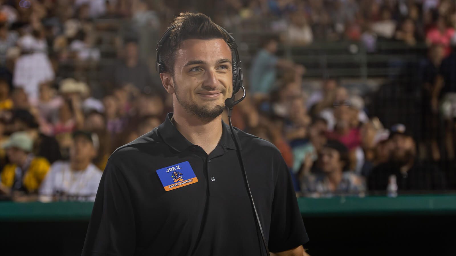 Full Sail grad Joe Zollo wears a headset on the field as Show Coordinator for the Savannah Bananas, with fans filling the stands behind him.