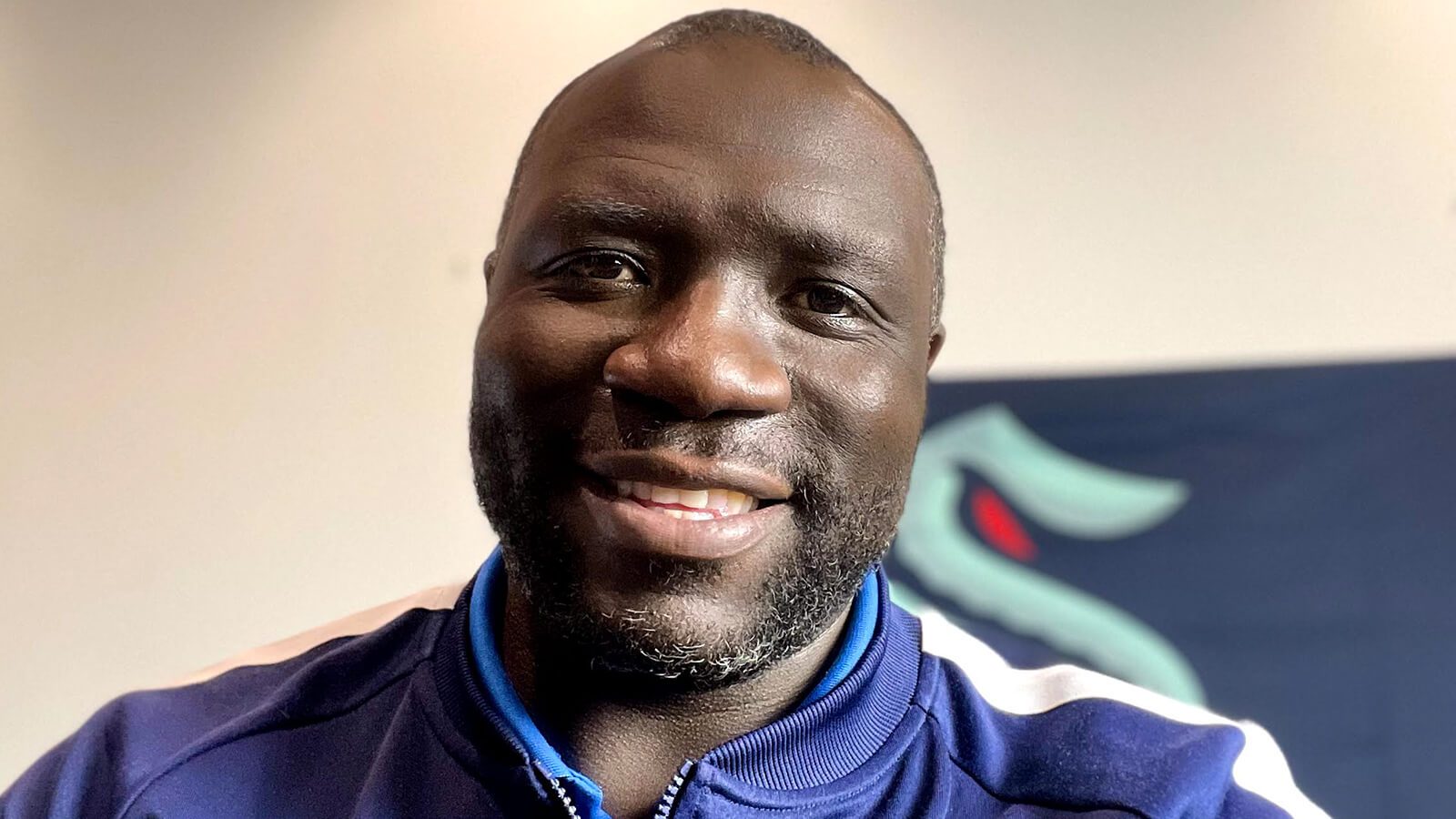 Grad Lamont Buford sports a blue jacket with a Seattle Kraken logo while smiling and holding a camera.