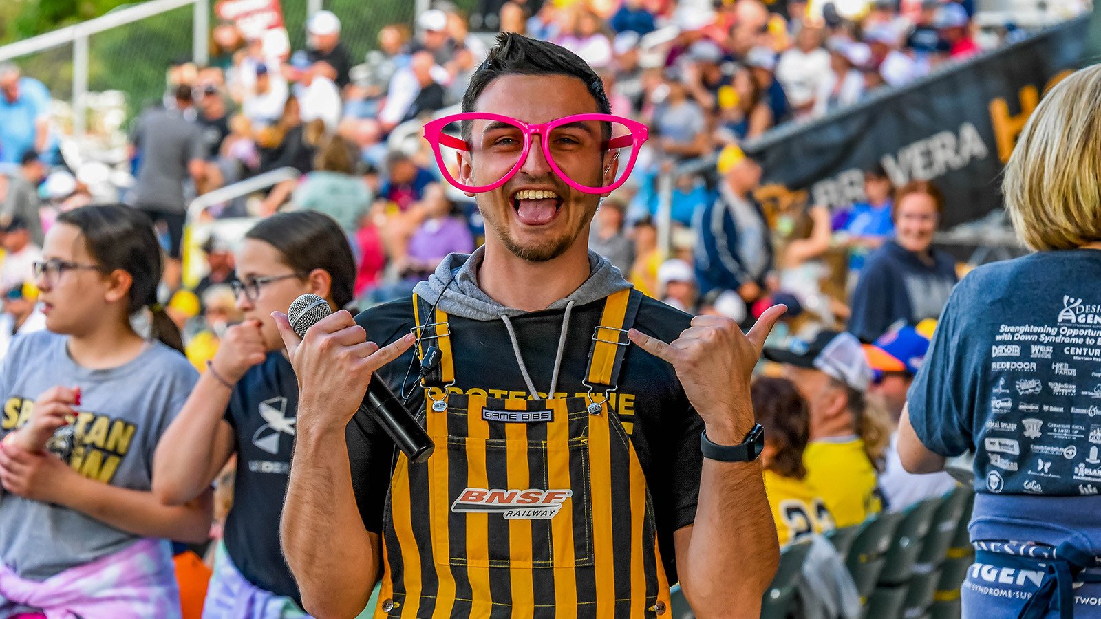 A man standing and looking excited while holding a microphone in oversized pink glasses and yellow and black striped overalls in the foreground of a large crowd at a stadium.